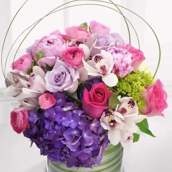 Low round arrangement of pink and lavender blooms with orchids and purple hydrangea in a glass vase