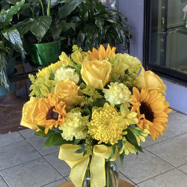 Yellow bouquet of roses, sunflowers, and mums in a glass vase with a ribbon