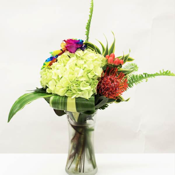 Modern bouquet of green hydrangeas, rainbow roses, and orange protea in a clear glass vase.