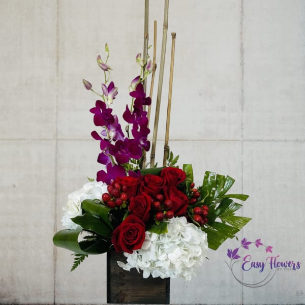 Tall floral arrangement with red roses, white hydrangeas, and purple orchids in a dark vase