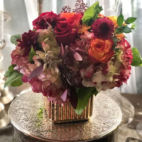 Compact bouquet of red, orange, and pink flowers in a gold vase