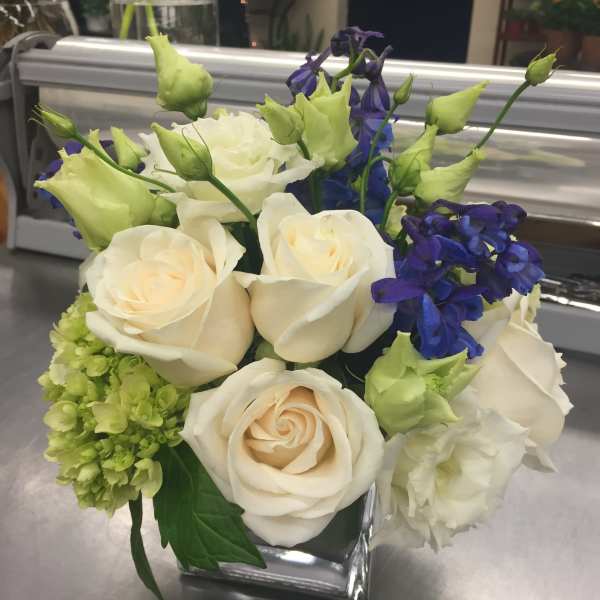 Cream roses and blue flowers arranged in a square glass vase