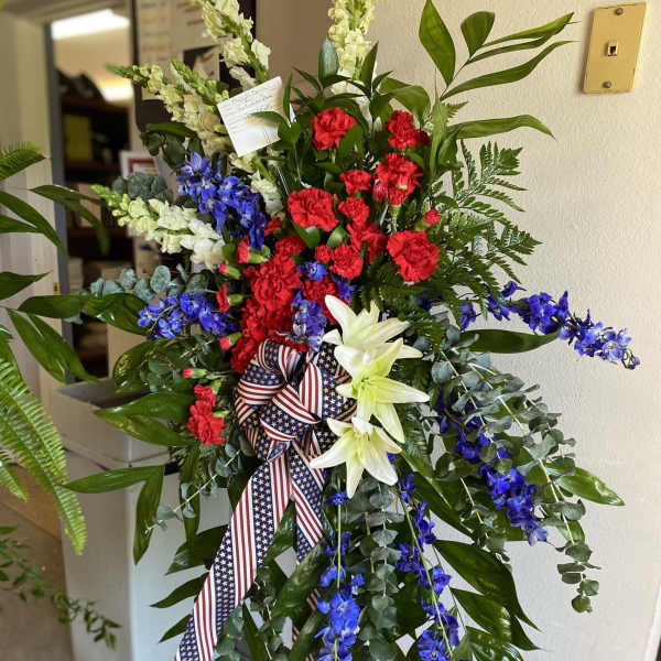 Patriotic standing spray with red carnations, white lilies, and blue blooms on an easel with flag ribbon.