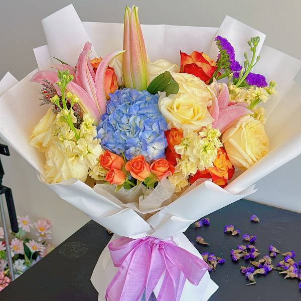 Bouquet of roses, lilies, and blue hydrangea wrapped in white paper with a purple ribbon