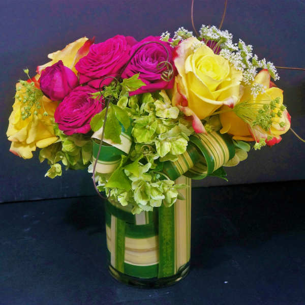 Compact arrangement of yellow and hot pink roses with green hydrangeas in a striped glass vase