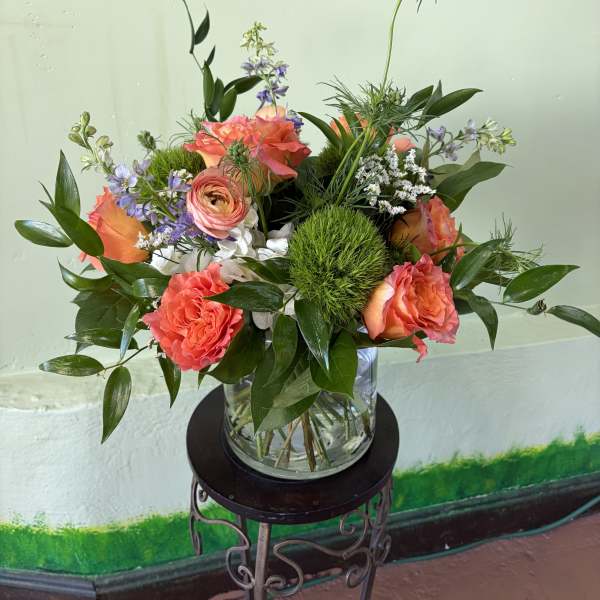 Coral roses, ranunculus, and green dianthus in a clear glass vase on a metal stand