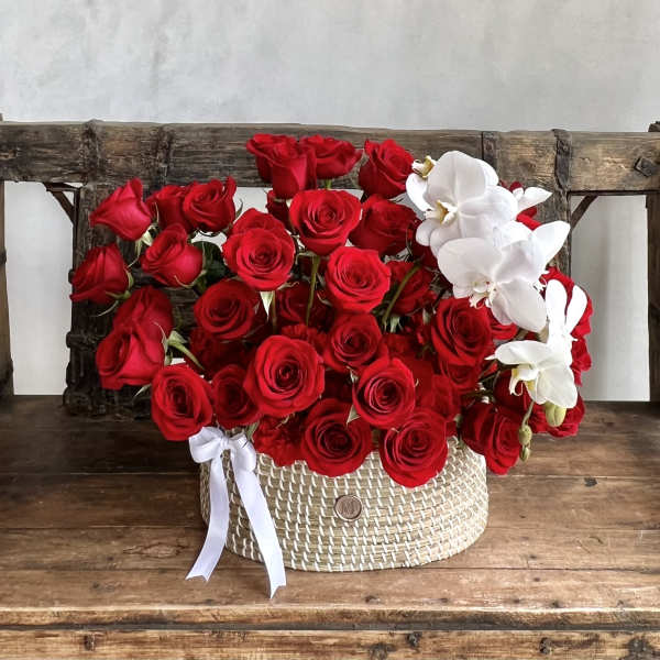 Red roses and white orchids arranged in a woven basket with a white ribbon