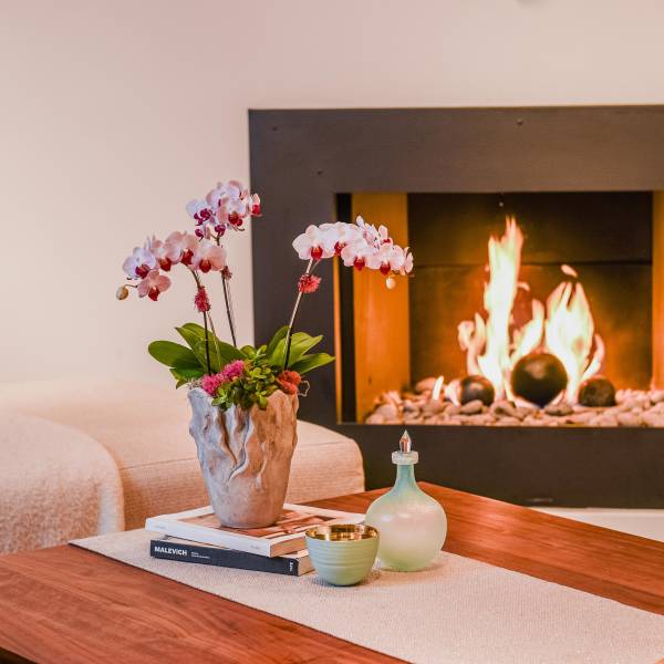 Potted orchids in a textured vase on a coffee table beside candles and a glass bottle.