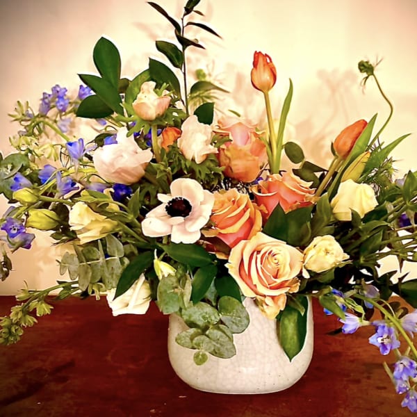 Low arrangement of peach roses, white anemones, and blue flowers in a white ceramic vase