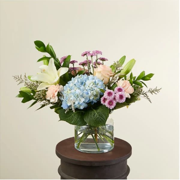 Pastel arrangement with blue hydrangea, white lilies, pink roses and daisies in a clear glass vase.