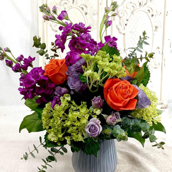 Vibrant arrangement of orange roses, purple stock, and green hydrangeas in a ribbed gray vase
