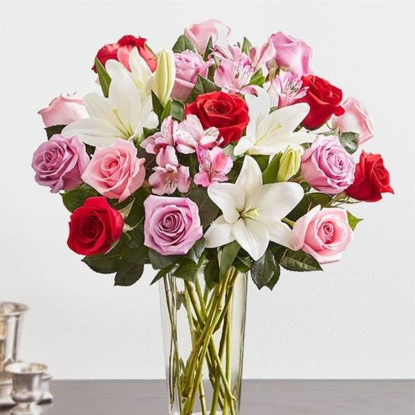 Arrangement of red, pink and lavender roses with white lilies in a tall clear glass vase