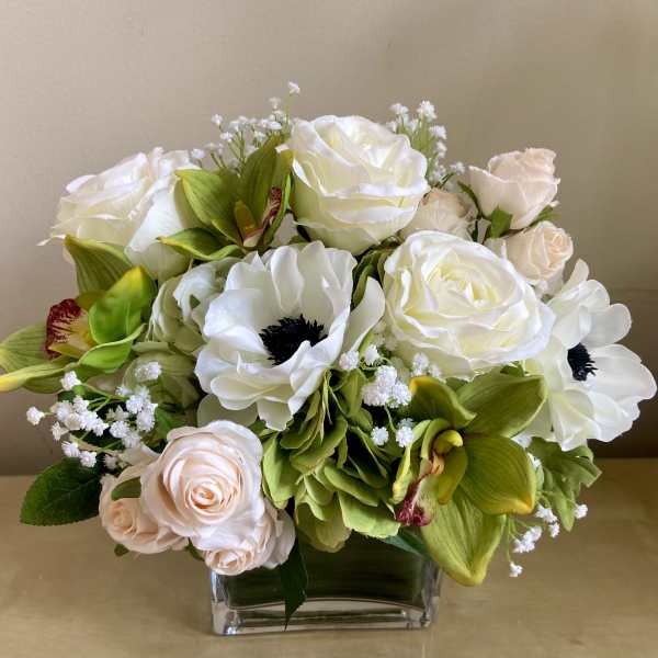Low arrangement of white and blush roses with white anemones and green orchids in a clear glass vase.