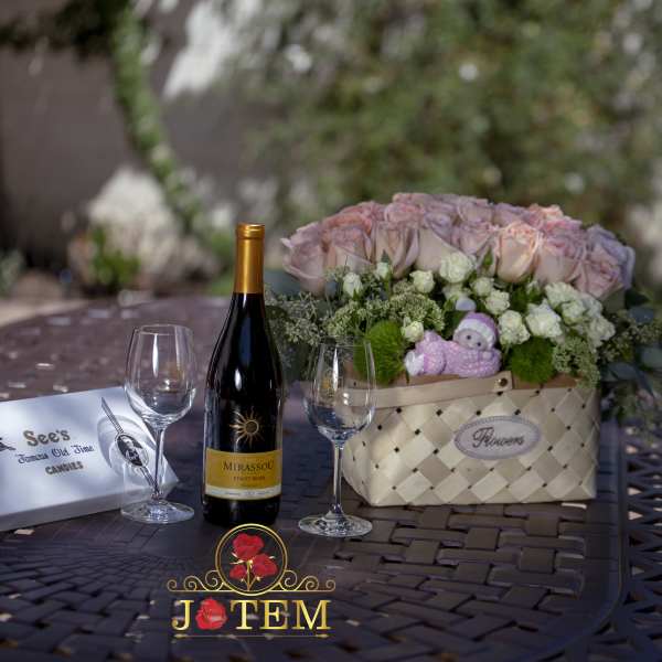 Pink rose arrangement in a woven basket with a small plush toy and wine bottle nearby