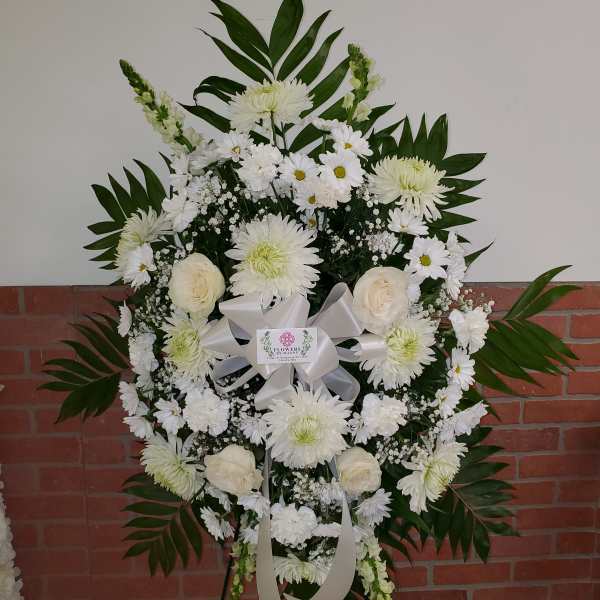Standing white funeral spray of roses, daisies, and mums with a white ribbon on an easel.