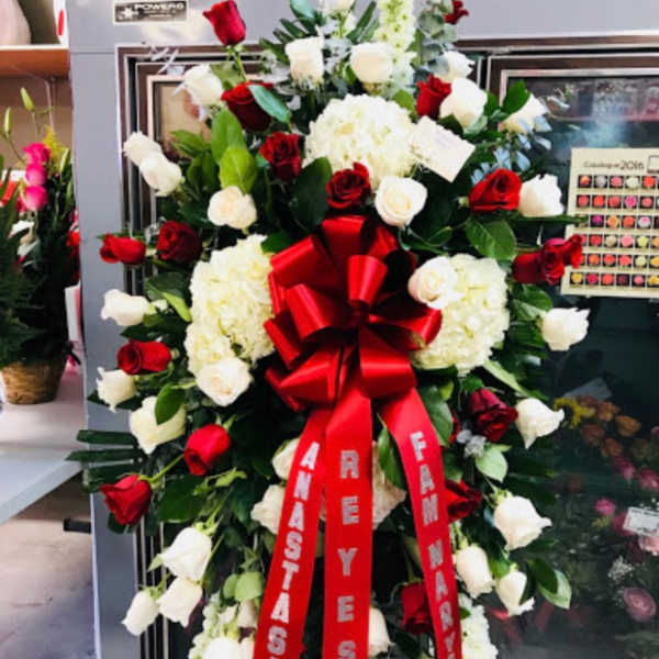 Tall standing spray of red and white roses with white hydrangeas and a large red bow on an easel.