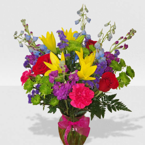 Bright mixed bouquet of yellow lilies, red roses, pink carnations, and purple blooms in a glass vase with pink bow