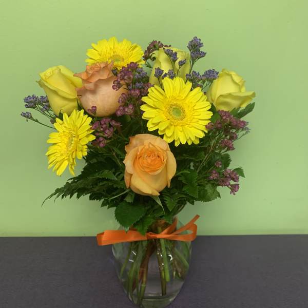 Yellow roses and gerbera daisies in a glass vase with an orange ribbon