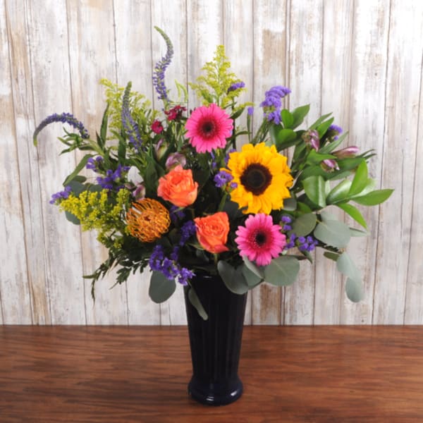 Tall mixed arrangement with sunflowers, pink gerbera daisies, and orange roses in a dark vase