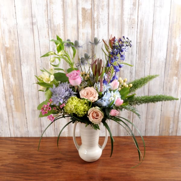 Tall mixed bouquet of pink roses, tulips, hydrangeas, and purple blooms in a white ceramic pitcher vase