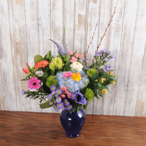 Colorful mixed flower arrangement in a blue vase with blue hydrangea, pink gerberas, and purple accents.