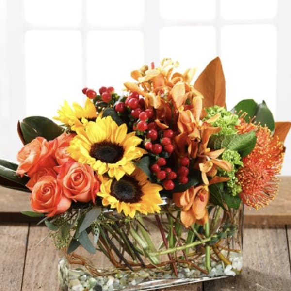 Low rectangular arrangement of sunflowers, orange roses, orchids, and berries in a clear glass vase.