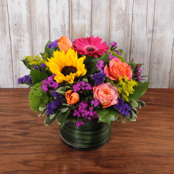 Low mixed bouquet with sunflower, pink gerbera, roses and purple flowers in a glass cylinder vase