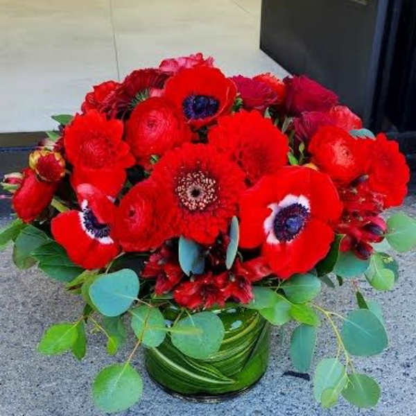 Low arrangement of vivid red flowers with dark centers in a leaf-wrapped glass vase