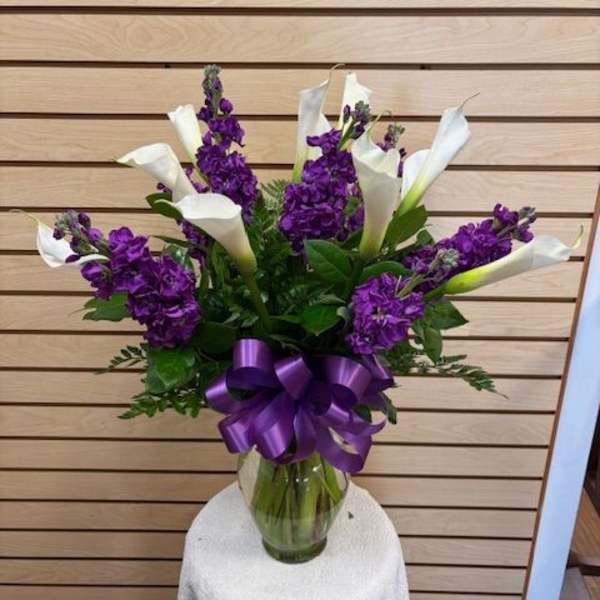 Purple flowers and white calla lilies in a glass vase with a purple ribbon