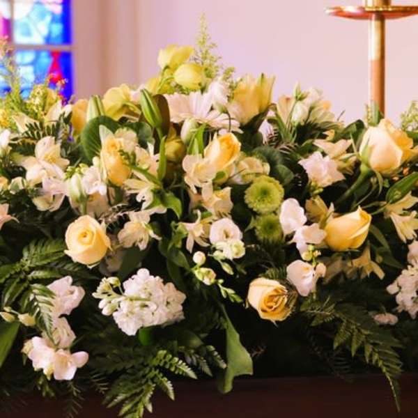 Long arrangement of white and pale yellow roses, lilies, and daisies on a casket in a church setting