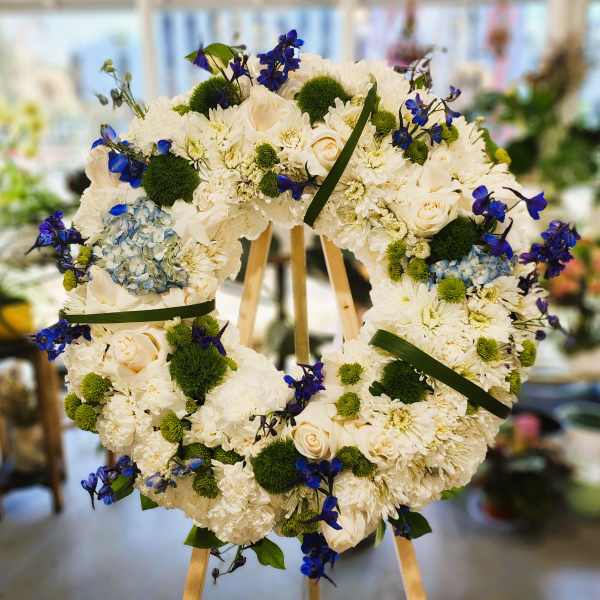 Heart-shaped floral wreath with white blooms, blue accents, and green pom-poms on an easel