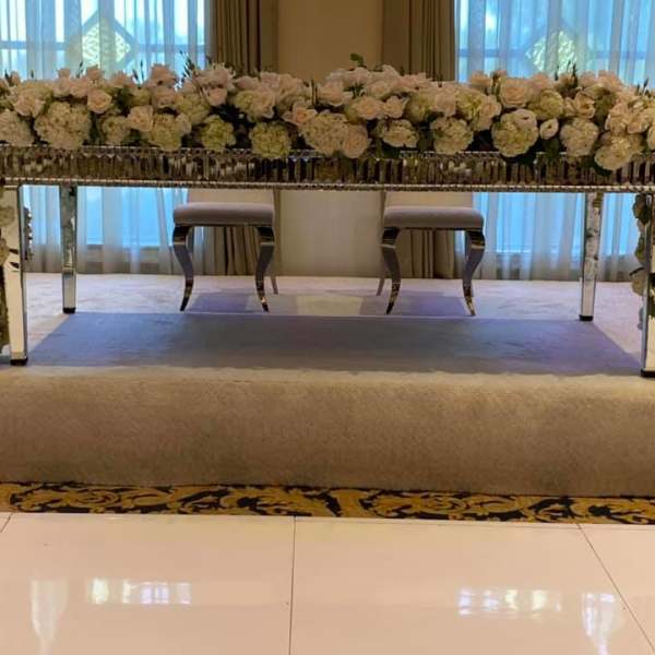 Long mirrored table wrapped in dense white and blush floral garlands on a stage.