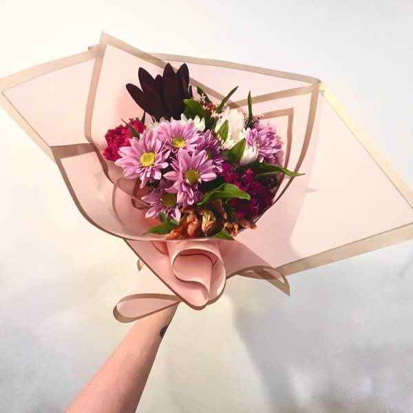 Handheld bouquet of pink and purple daisies wrapped in blush paper