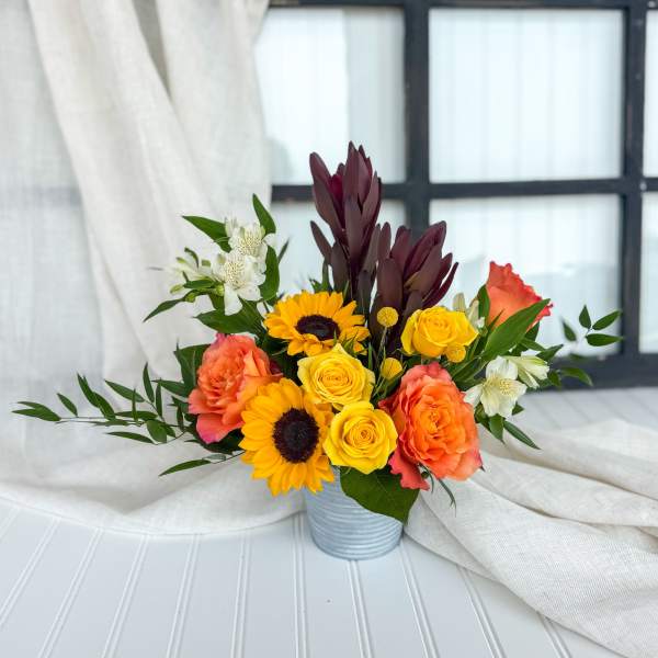 Mixed bouquet of yellow roses, sunflowers, and coral blooms in a metal container