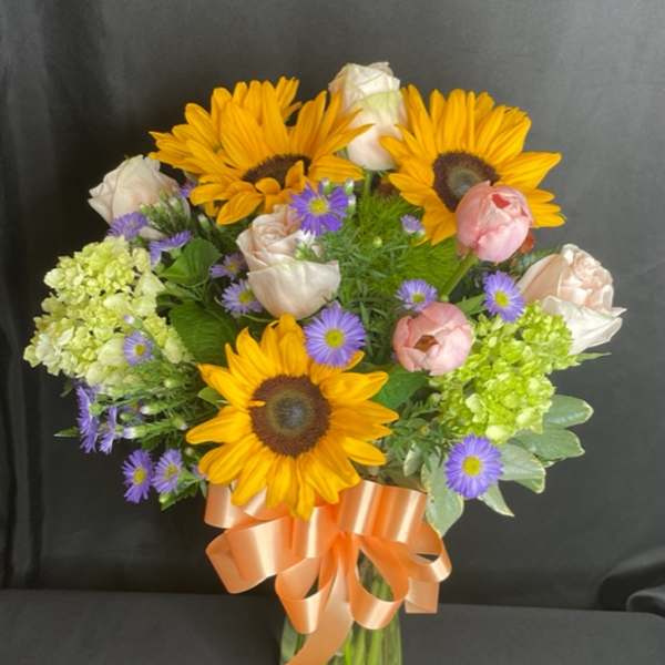 Bouquet of sunflowers, roses, and purple daisies in a glass vase with a peach ribbon