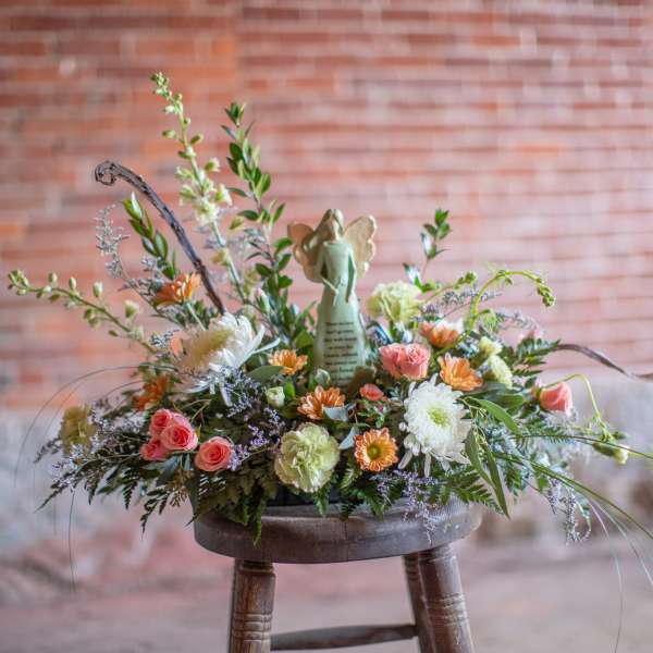 Low pastel arrangement of roses, mums, and daisies with an angel figurine in the center on a rustic stool