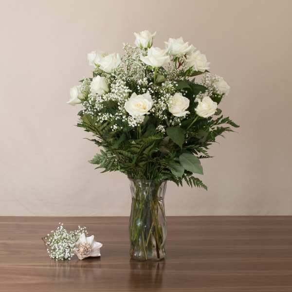 Tall arrangement of white roses and baby's breath in a clear glass vase with a small matching corsage on the table