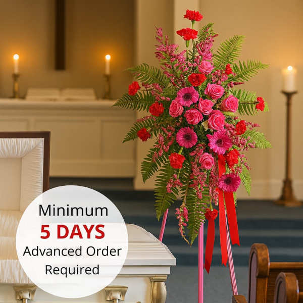 Bright pink and red standing spray on an easel beside a white casket in a chapel.