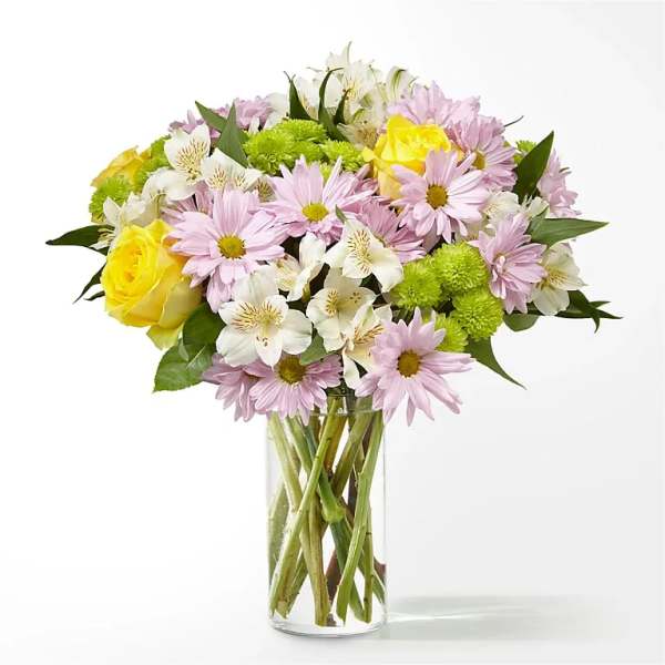 Mixed bouquet of pink daisies, yellow roses, and white flowers in a glass vase