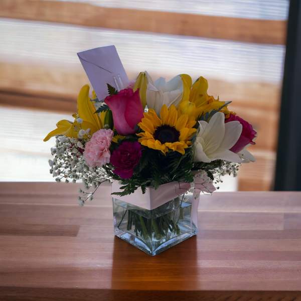 Mixed bouquet in a clear square vase with sunflowers, lilies, and roses