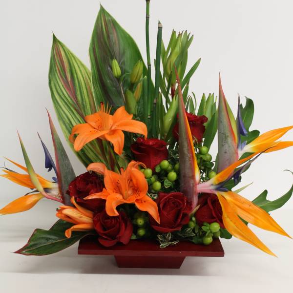 Tropical arrangement with orange lilies, red roses, and birds of paradise in a low red container