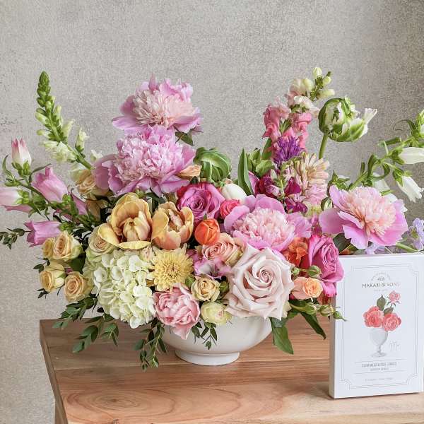 Large pastel flower arrangement in a white bowl with a cookie box beside it.