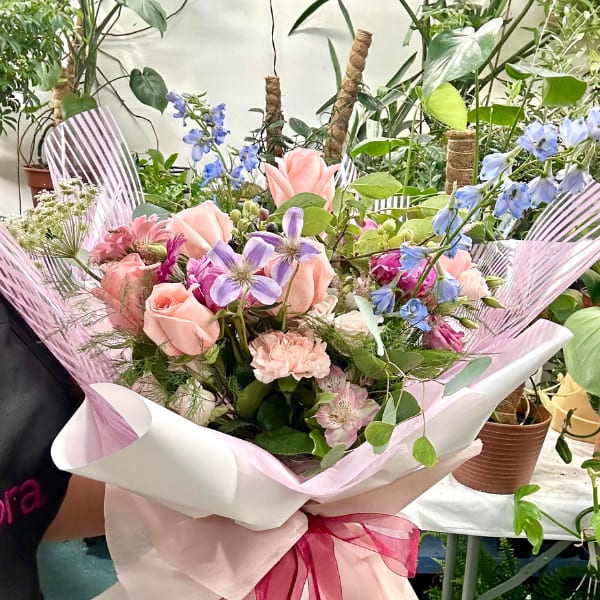 Hand-tied bouquet of peach roses and pastel mixed flowers wrapped in white and pink paper with a magenta ribbon