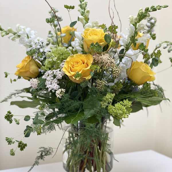 Tall arrangement of yellow roses, white snapdragons, and green accents in a clear glass vase