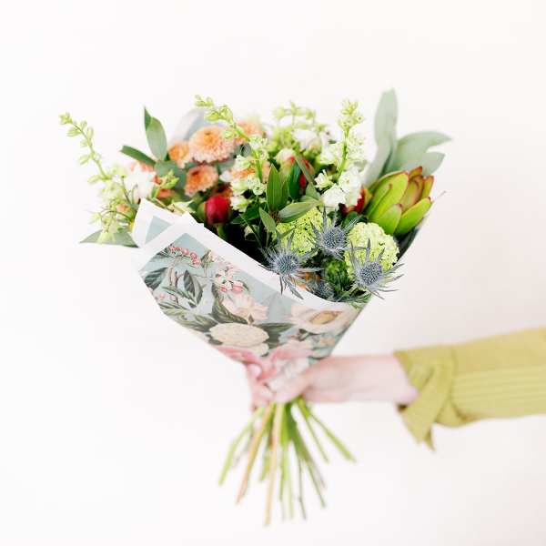 Handheld bouquet with peach, green, and blue flowers in floral wrap