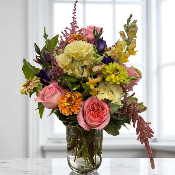 Mixed bouquet of pink roses, yellow carnations, and purple blooms in a glass vase