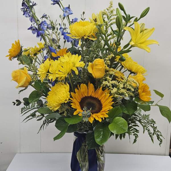 Tall bouquet of yellow and blue flowers in a glass vase with a dark ribbon