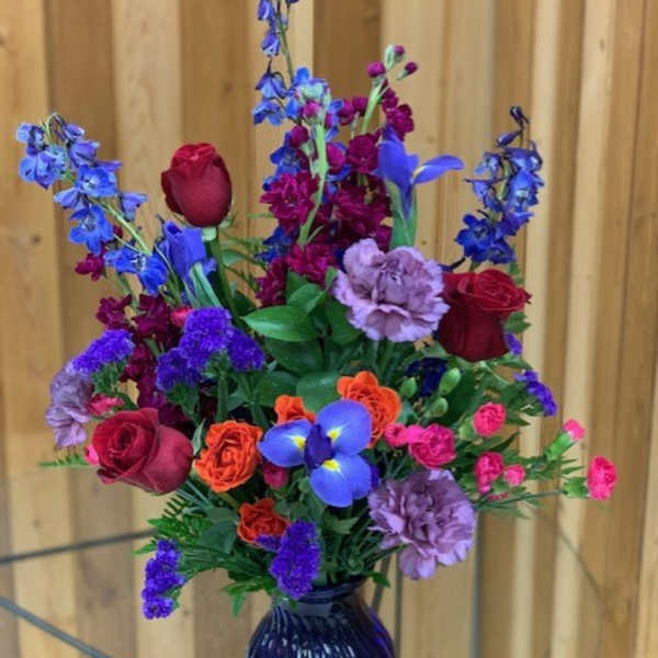 Tall mixed arrangement with red roses, purple delphinium and carnations in a blue glass vase