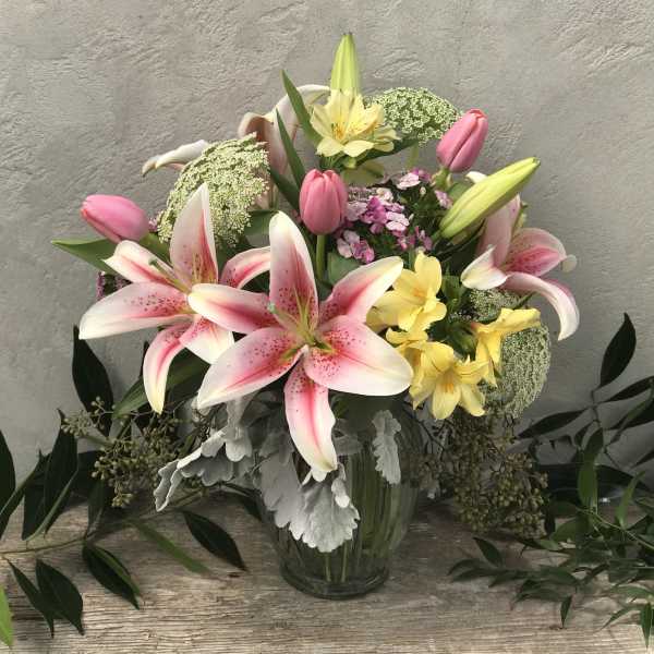 Pink lilies, tulips, and yellow flowers arranged in a clear glass vase against a neutral background