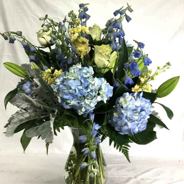 Tall arrangement of blue hydrangeas, white roses, lilies, and yellow flowers in a clear glass vase.